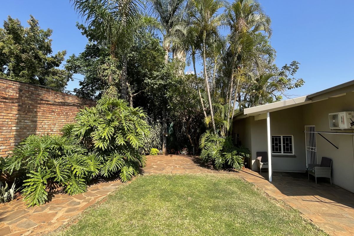 A private rear garden featuring a manicured lawn, stone paved pathways, mature palm trees, and a brick boundary wall.