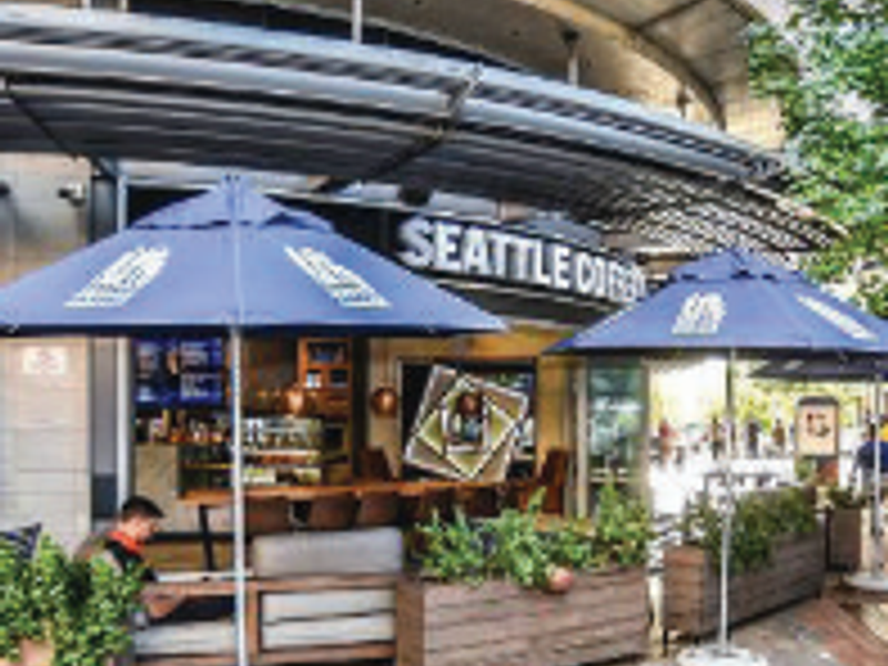 Outdoor seating area for a coffee shop featuring a curved concrete canopy, paved flooring, and integrated wooden planters.
