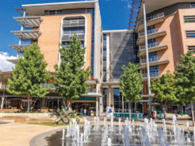A modern multi-story office building featuring a brick and glass facade, private balconies, and a prominent water fountain in the landscaped courtyard.