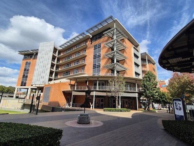 A modern multi-story office building featuring a brick facade, integrated metal balconies, and a spacious paved entrance courtyard.