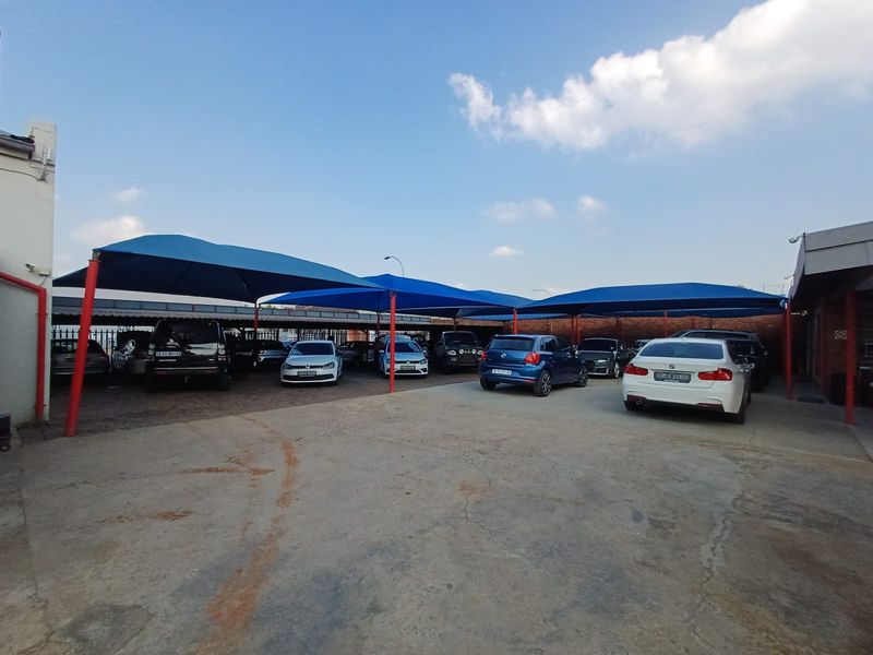 Commercial parking area featuring extensive blue shade cloth structures supported by red steel poles, providing covered parking bays for multiple vehicles.
