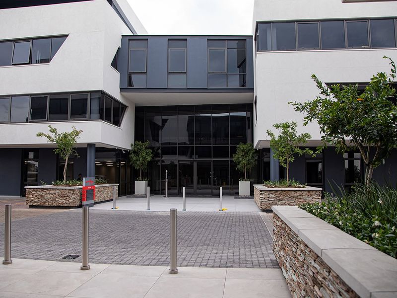 Modern office building entrance featuring a glass facade, stone-clad planters, and a paved approach with integrated security bollards.