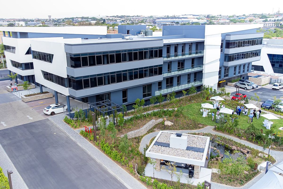 Aerial view of a modern multi-story office complex featuring contemporary glass architecture, landscaped gardens, a water feature, and paved parking areas.
