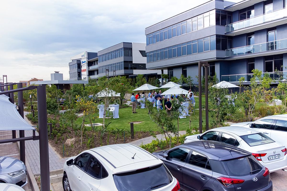 A well-maintained landscaped garden area situated between modern multi-story office buildings, featuring paved pathways and integrated shade structures for outdoor use.