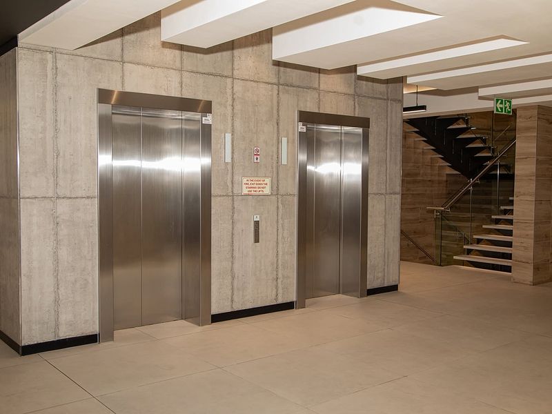 Modern office building lobby featuring dual stainless steel elevators, tiled flooring, and a contemporary staircase with glass balustrades.
