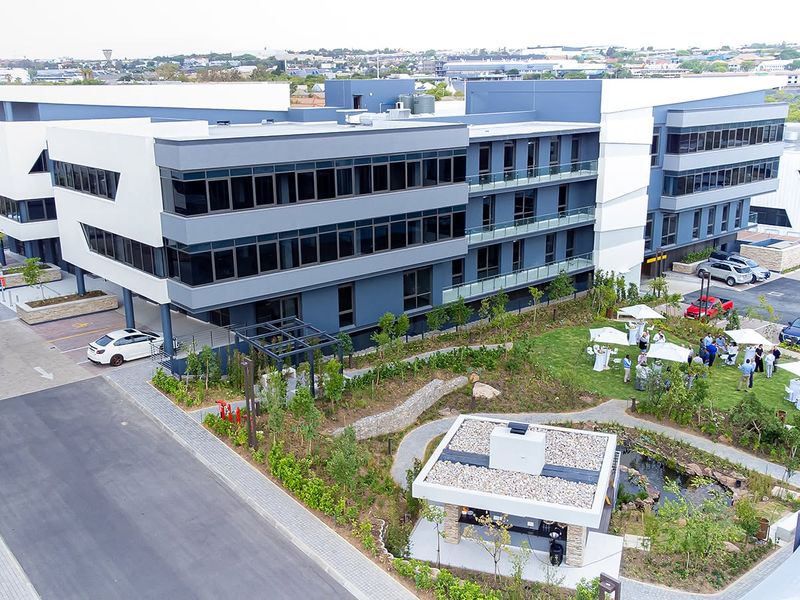Aerial view of a modern multi-story office complex featuring contemporary glass architecture, landscaped gardens, a water feature, and paved parking areas.