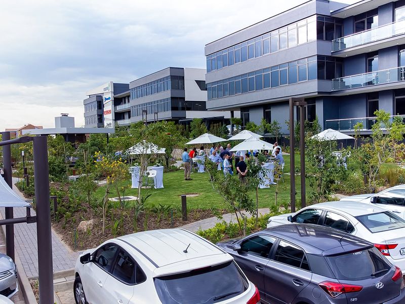 A well-maintained landscaped garden area situated between modern multi-story office buildings, featuring paved pathways and integrated shade structures for outdoor use.