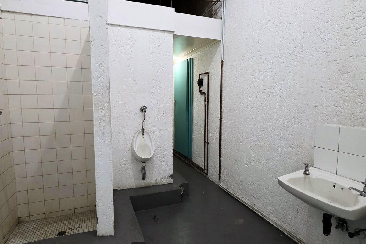Industrial-style communal bathroom featuring a tiled shower enclosure, wall-mounted urinal, and dual basins with exposed plumbing fixtures.