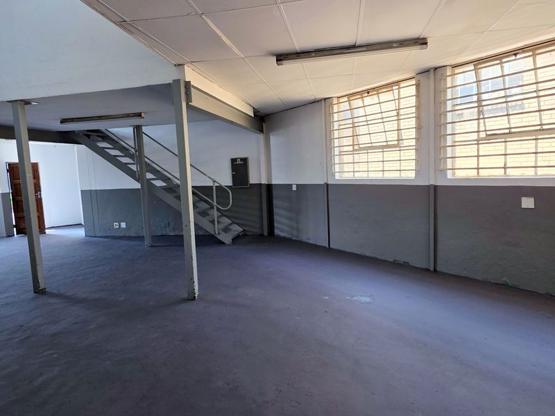 Spacious open-plan industrial unit featuring a steel staircase, mezzanine potential, secure window bars, and durable painted concrete flooring throughout.