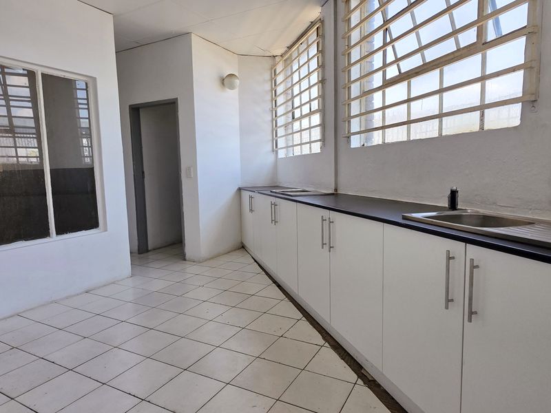 A functional industrial kitchen space featuring white built-in cabinetry, a dark laminate countertop, stainless steel sink, and tiled flooring with security-barred windows.