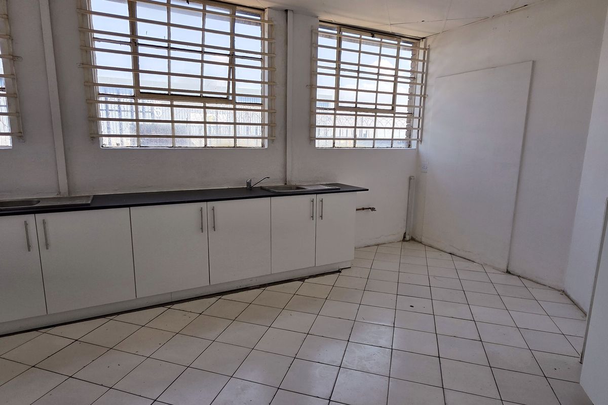 A functional industrial kitchen space featuring white built-in cabinetry, dark countertops, stainless steel sink, and tiled flooring with security bars on the windows.