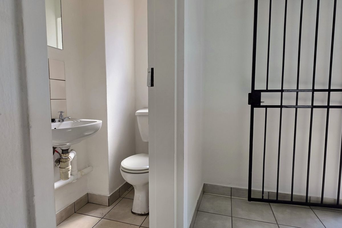 A functional bathroom featuring a pedestal washbasin and toilet, with tiled flooring and direct access through a security gate.
