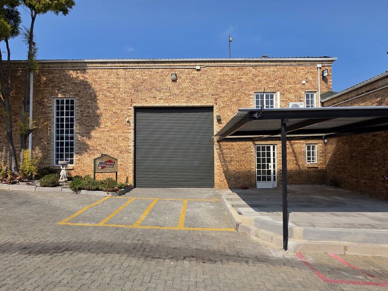 Industrial property exterior featuring a brick facade, large roller shutter door, paved parking area, and a covered carport.