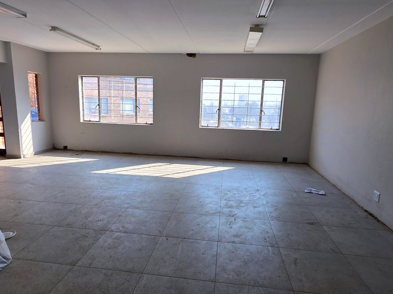 An empty, open-plan industrial office space featuring tiled flooring, fluorescent lighting, and large steel-framed windows, currently awaiting renovation or fit-out.