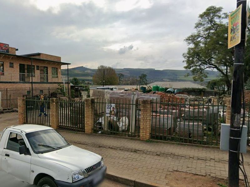 Street-level view of a vacant commercial plot featuring brick and metal perimeter fencing, situated in an established neighborhood setting.