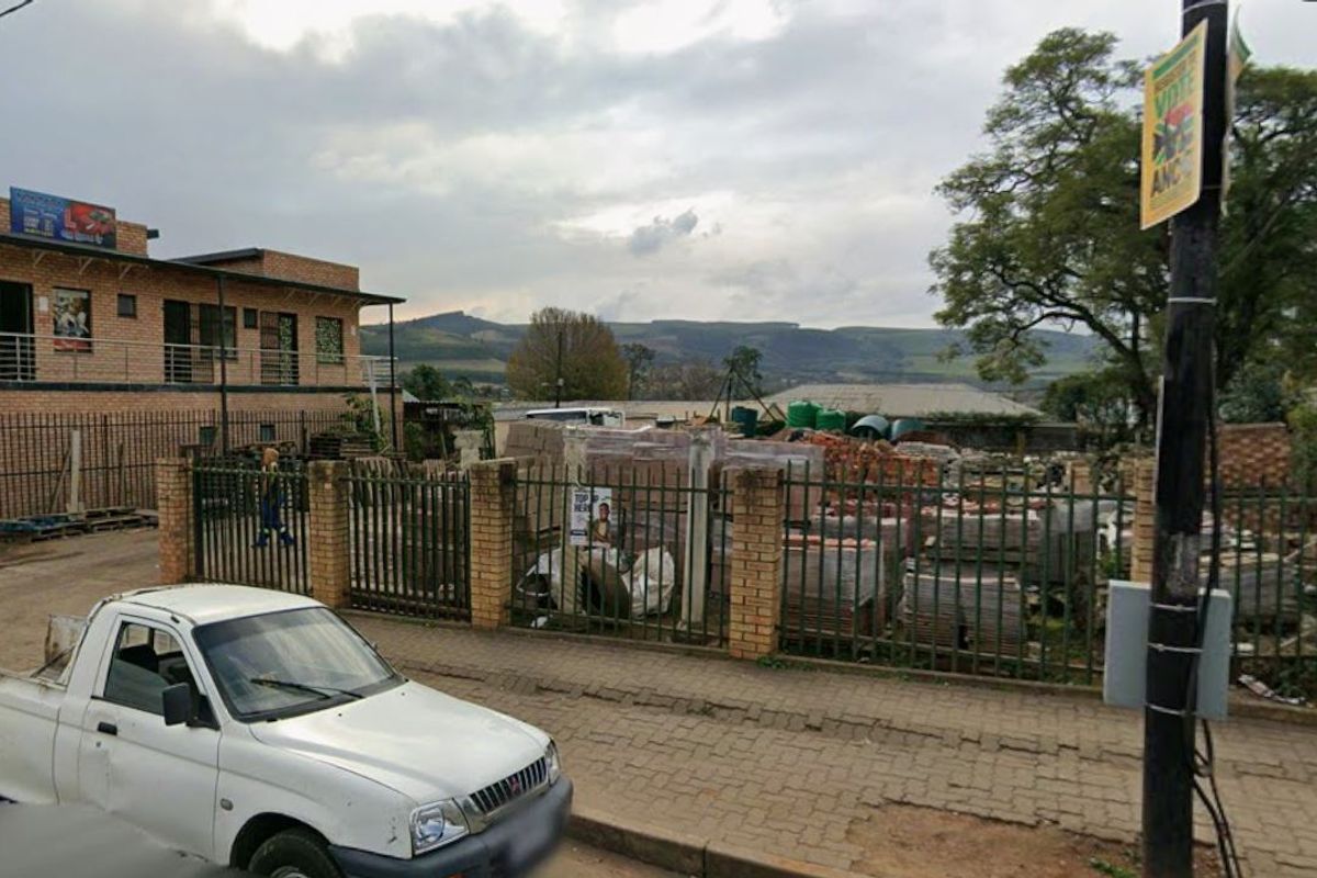 Street-level view of a vacant commercial plot featuring brick and metal perimeter fencing, situated in an established neighborhood setting.