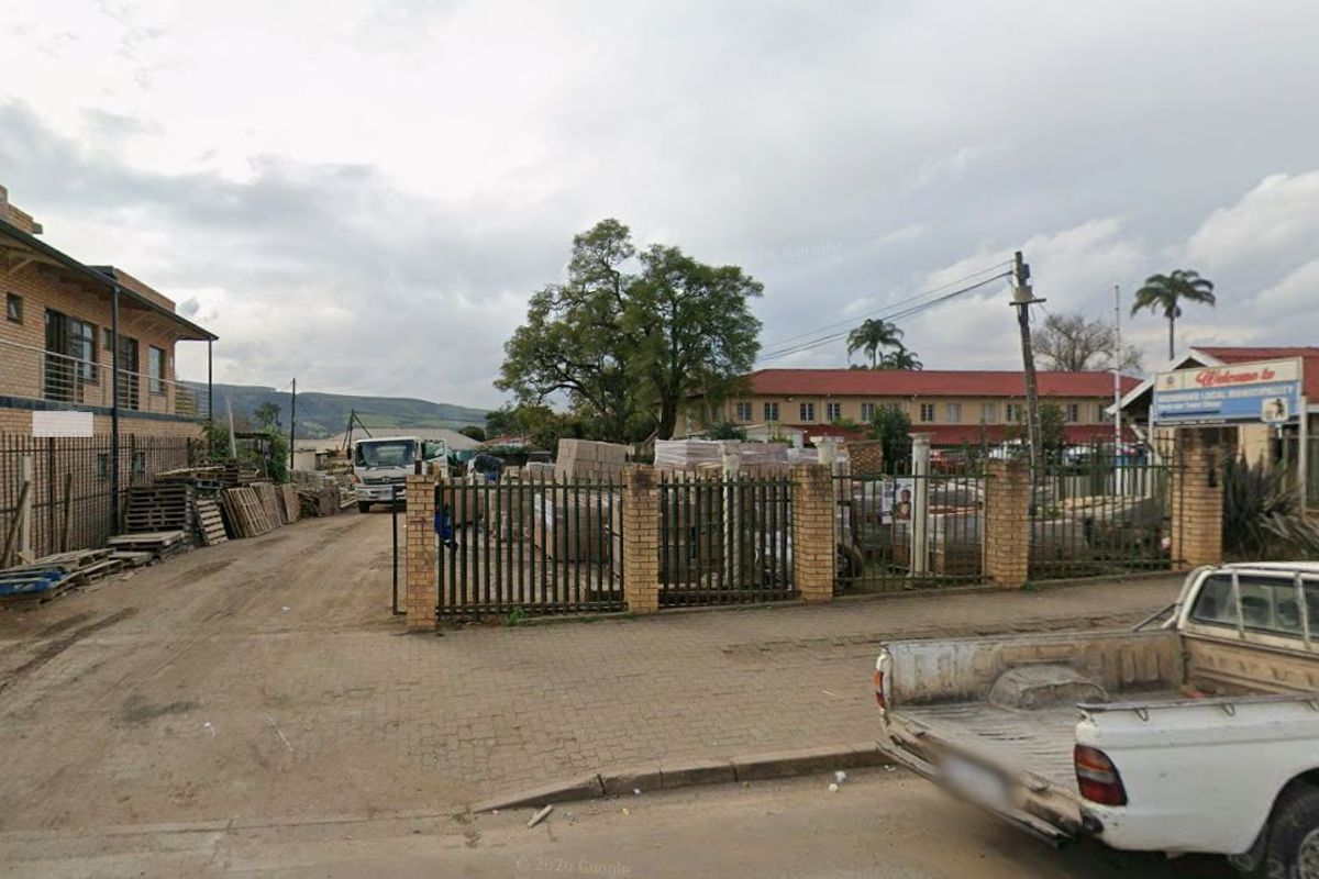 Street-level view of a commercial property featuring a paved driveway, brick perimeter wall, and metal security fencing.