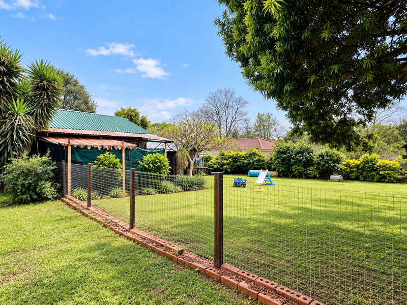 A spacious, well-maintained garden area featuring a wire mesh fence with a brick border, mature trees, and a small outbuilding with a metal roof.