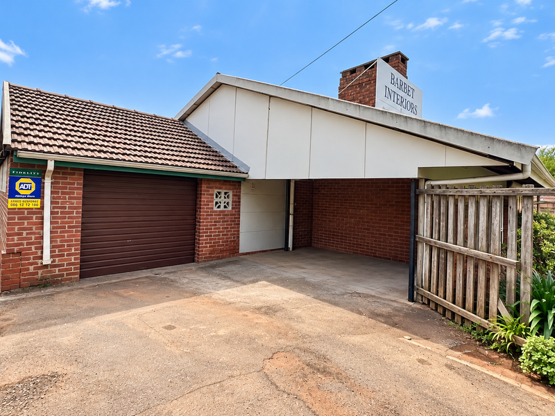 Exterior view of a business property featuring a brick garage, attached carport, and concrete driveway with perimeter timber fencing.