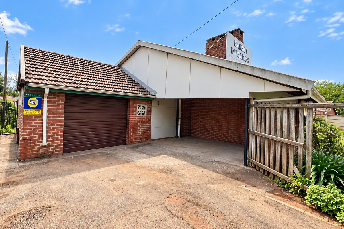 Exterior view of a business property featuring a brick garage, attached carport, and concrete driveway with perimeter timber fencing.