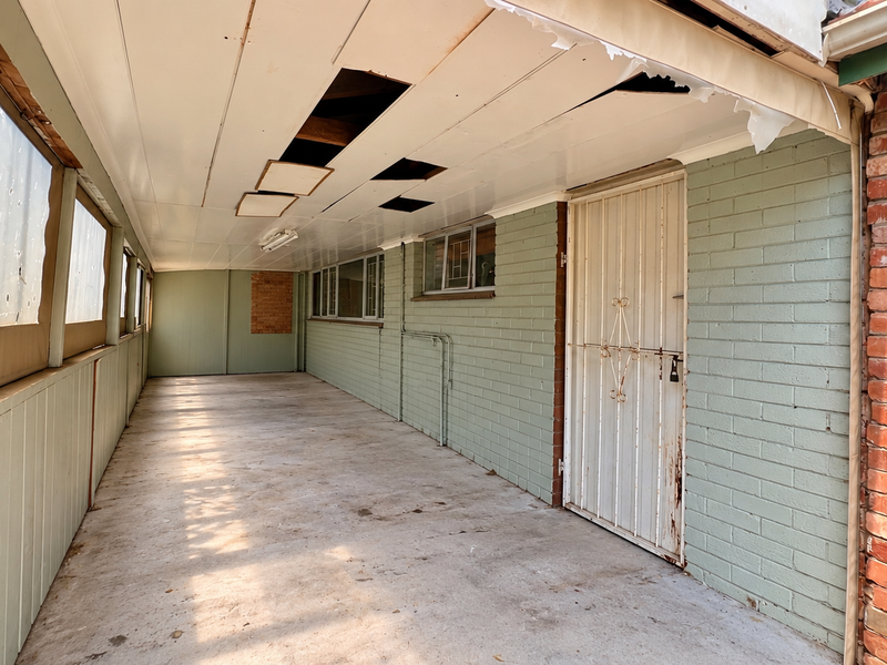 A covered outdoor patio area featuring painted brick walls, concrete flooring, and a security gate, currently requiring ceiling and screening repairs.