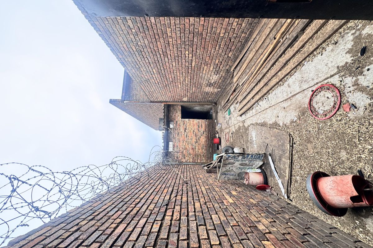 A narrow, enclosed outdoor utility passage featuring high brick walls topped with security razor wire and a concrete pathway.