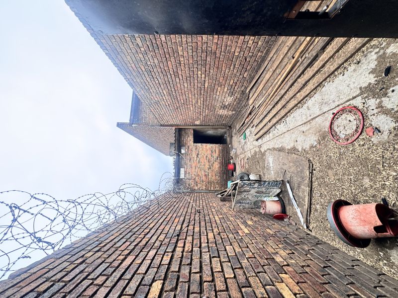 A narrow, enclosed outdoor utility passage featuring high brick walls topped with security razor wire and a concrete pathway.