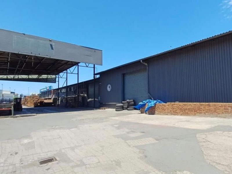 Industrial yard area featuring a large warehouse building with corrugated metal cladding, a roller shutter door, and a covered loading bay.
