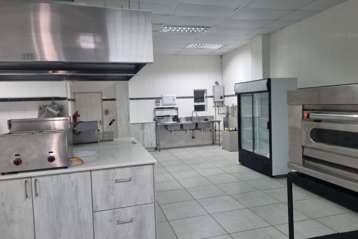 A well-equipped commercial kitchen featuring stainless steel prep stations, a large extractor hood, triple basin sink, and durable tiled finishes throughout.