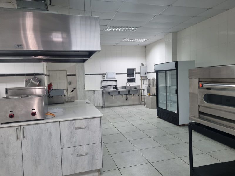 A well-equipped commercial kitchen featuring stainless steel prep stations, a large extractor hood, triple basin sink, and durable tiled finishes throughout.