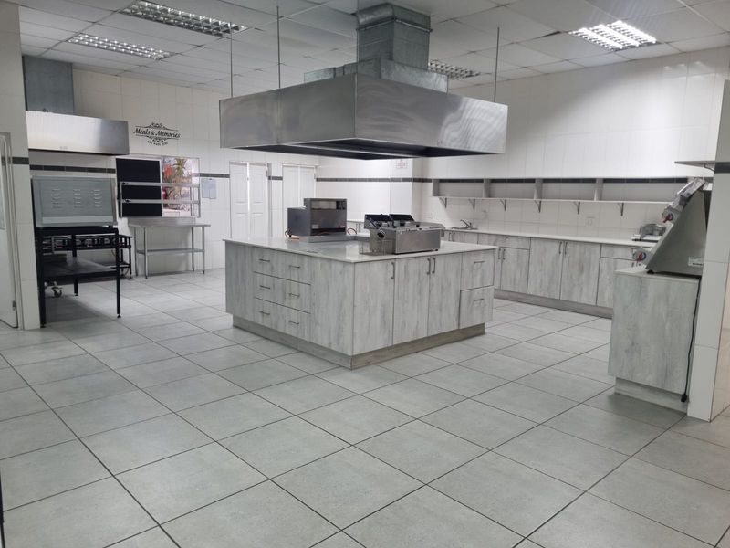 A spacious commercial kitchen featuring tiled surfaces, a large central island with stainless steel extractor hood, and ample built-in cabinetry and shelving.