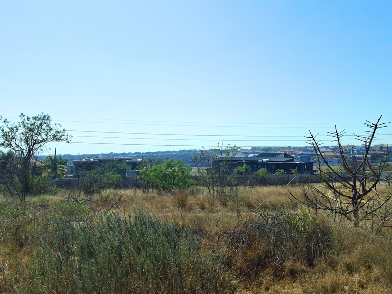 A view of a vacant commercial land plot featuring natural vegetation and overhead power lines with surrounding residential development in the distance.