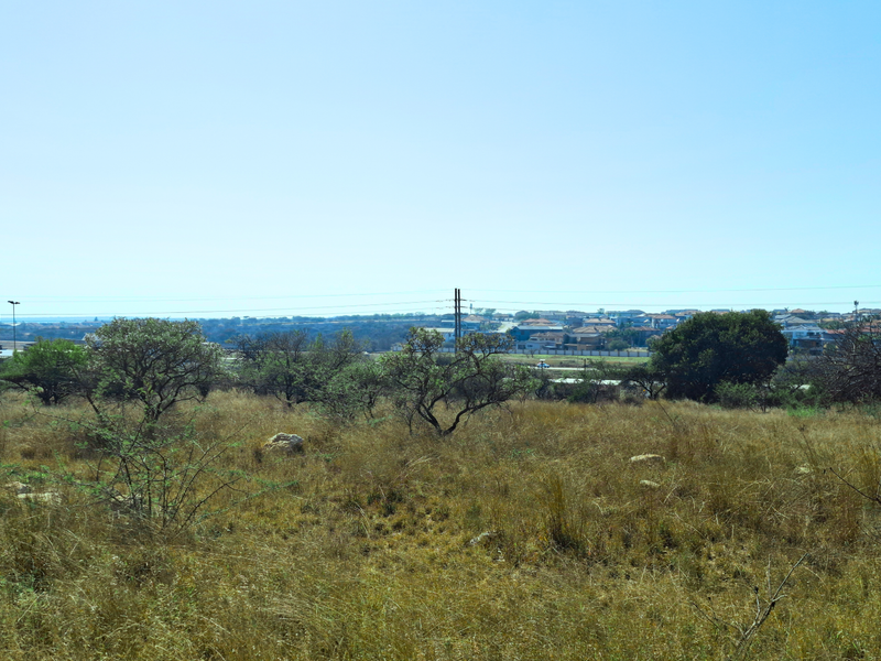 A wide-angle view of a vacant commercial land plot featuring natural vegetation and distant residential development under a clear sky.