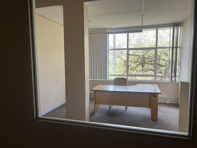 A functional office space featuring large windows with vertical blinds, carpeted flooring, and a glass partition wall, offering a bright and professional work environment.