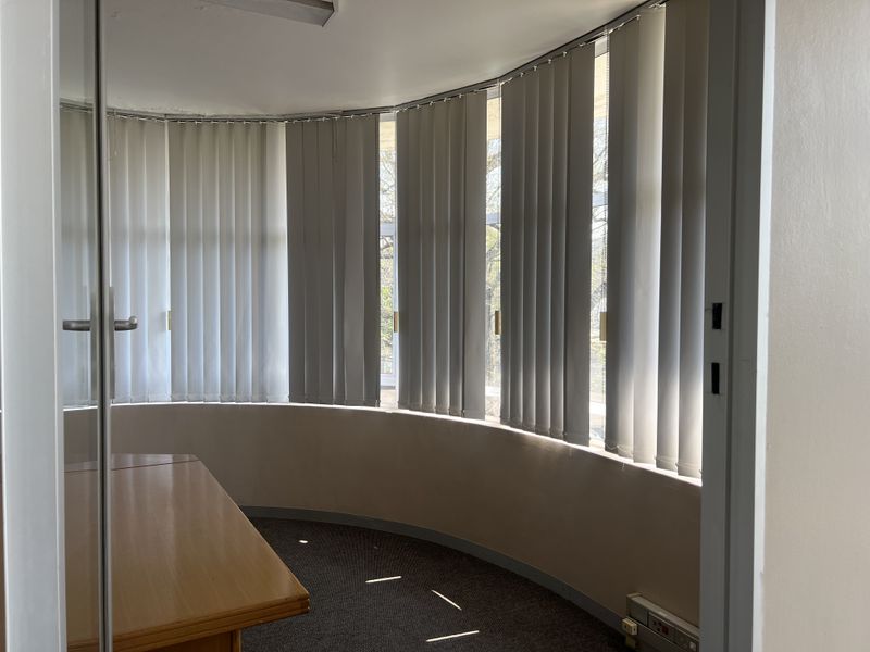 A curved office space featuring large windows with vertical blinds, carpeted flooring, and a built-in desk area.