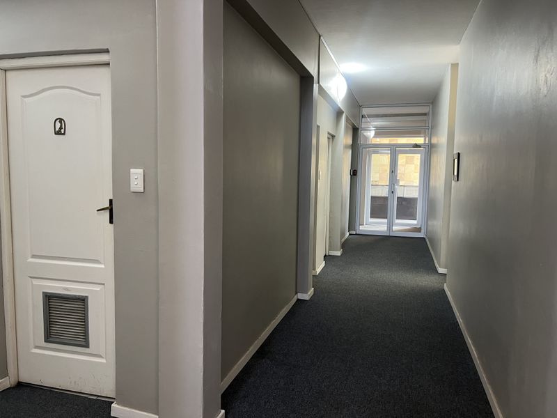 A clean, functional commercial hallway featuring grey carpeting, neutral walls, and a glass-panelled door providing natural light and access to the exterior.