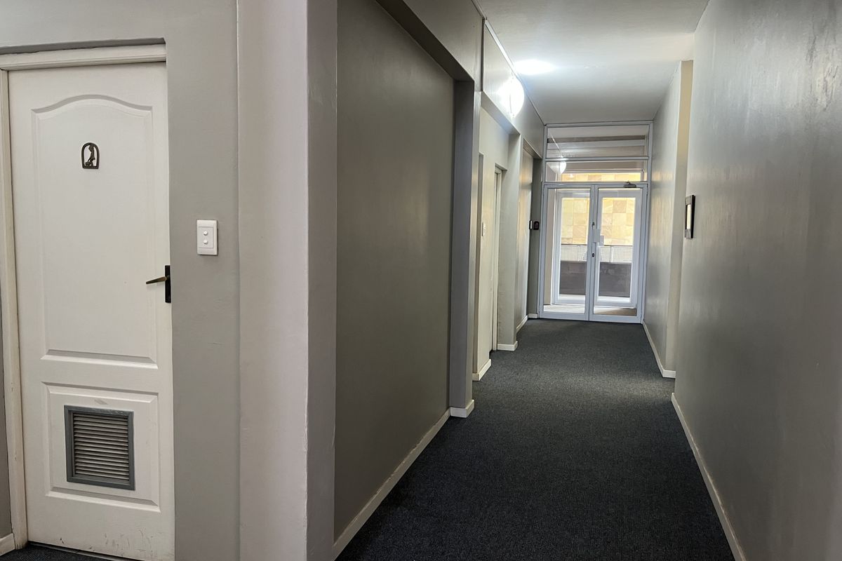 A clean, functional commercial hallway featuring grey carpeting, neutral walls, and a glass-panelled door providing natural light and access to the exterior.