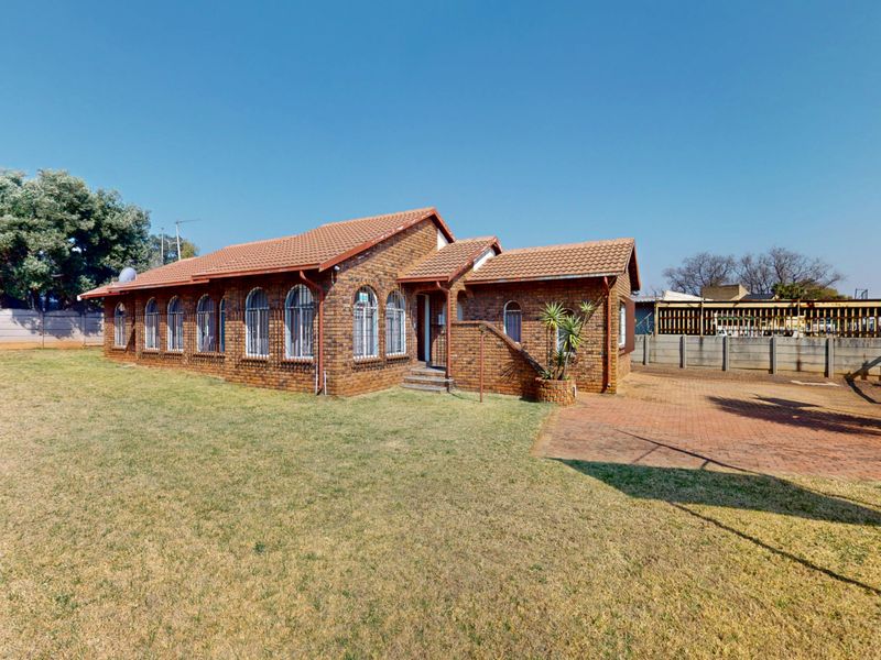 A rear view of a face brick building featuring arched windows with security bars, a tiled roof, and a spacious lawn with a paved driveway.
