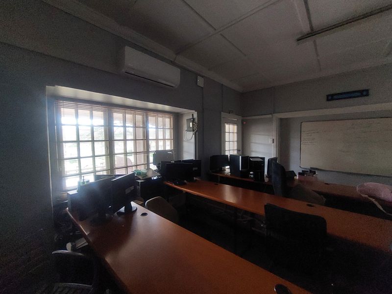 A functional commercial office space featuring built-in desks, wall-mounted air conditioning unit, and a whiteboard, suitable for a training or workspace environment.