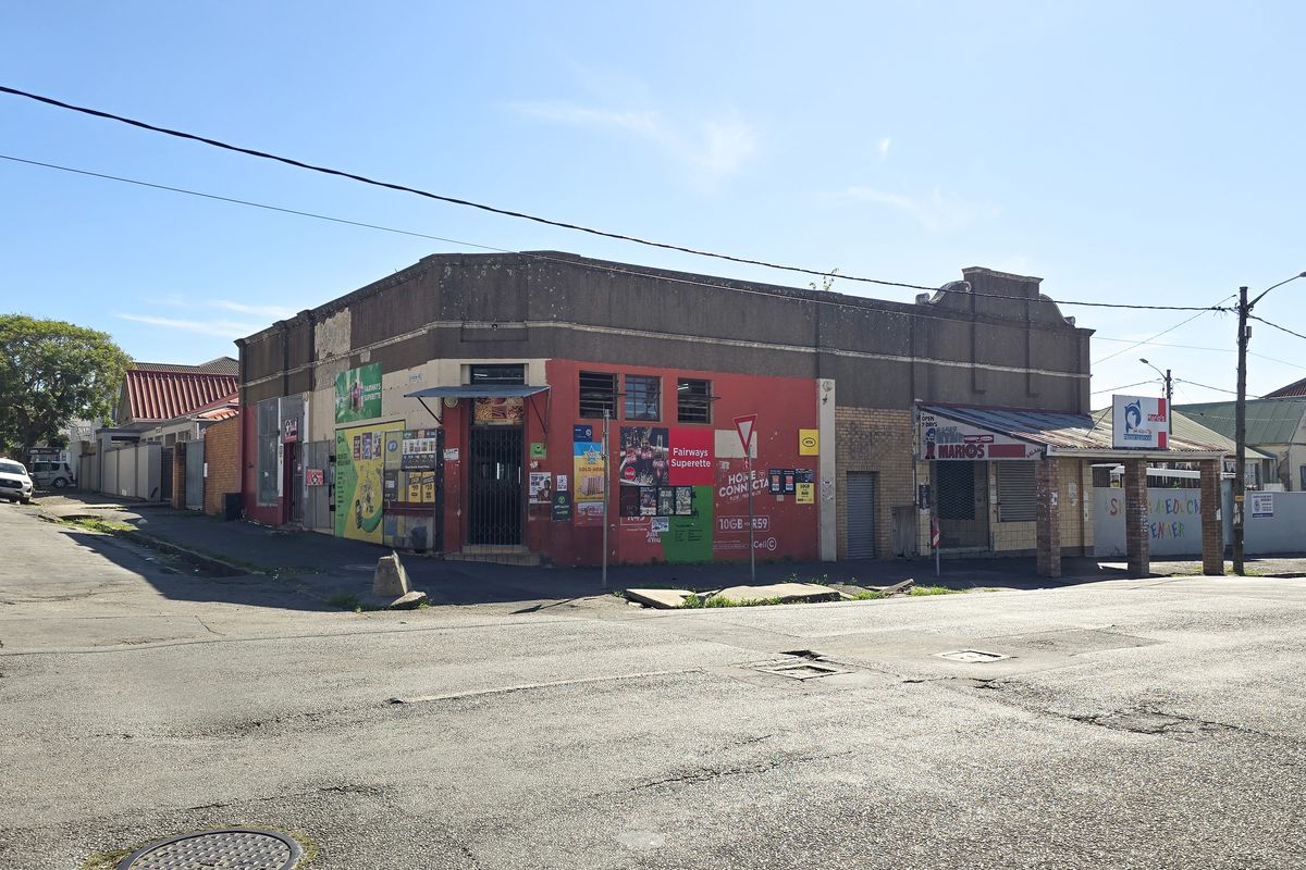 Street-level view of a commercial corner building featuring multiple storefronts with security gates and roller shutters in a high-traffic urban location.
