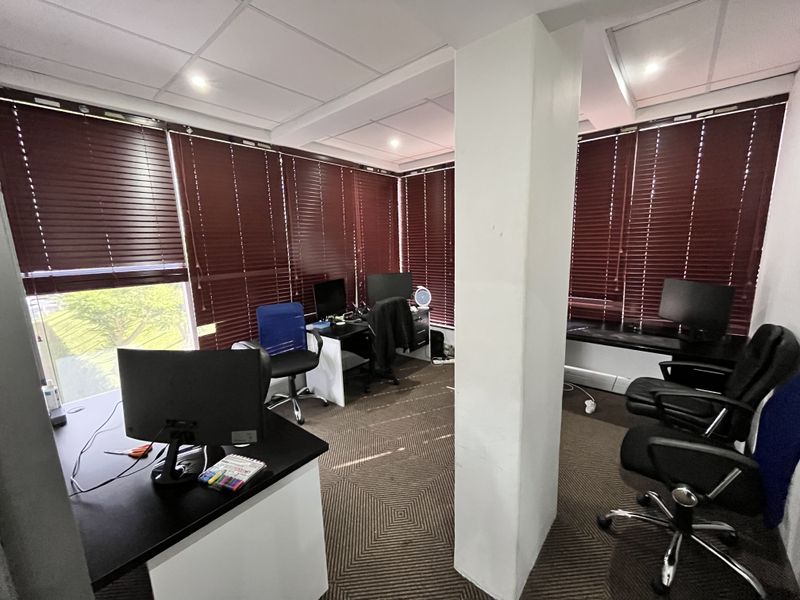 A functional office space featuring a central structural column, suspended ceiling tiles, and large windows fitted with dark red venetian blinds.
