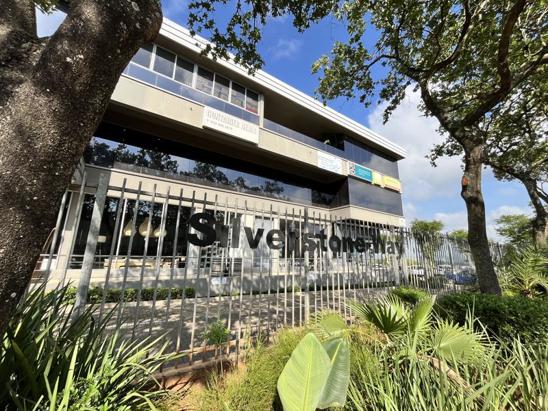 A modern multi-story office building facade featuring glass curtain walls, metal security fencing, and prominent signage, set within a landscaped commercial environment.
