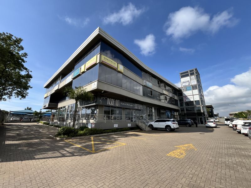 Modern multi-story commercial office building featuring glass curtain walls, paved parking area, and a prominent glass-enclosed stairwell tower.