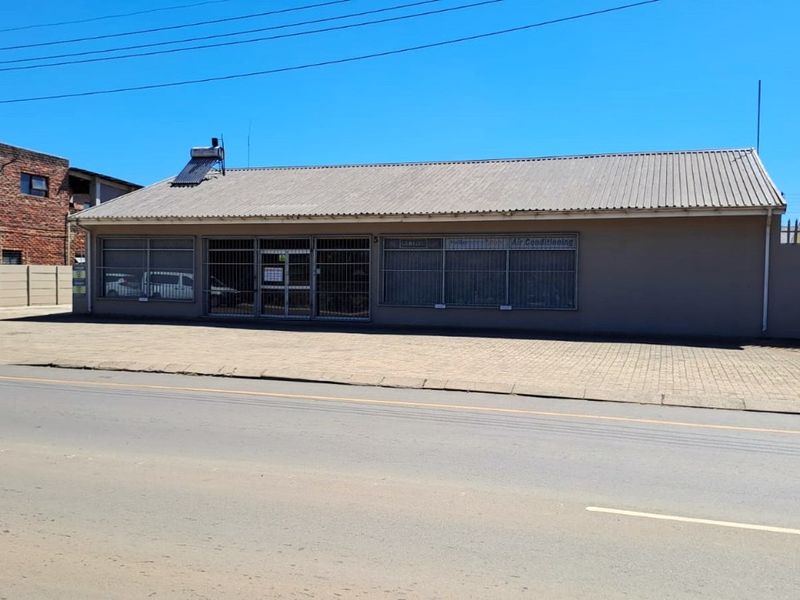 Street-facing commercial property featuring a paved frontage, large display windows with security bars, and a pitched roof with solar water heating.