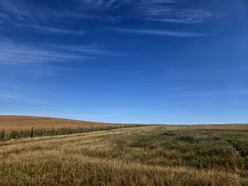 Expansive view of open farmland featuring rolling fields and perimeter wire fencing under a clear blue sky.