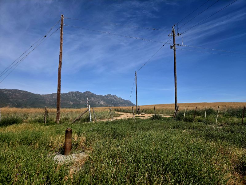 A wide landscape view of the farm property featuring open fields, wire fencing, and utility infrastructure against a mountain backdrop.