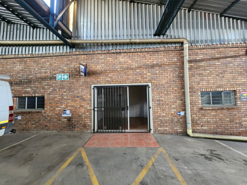 Industrial unit entrance featuring exposed brickwork, a secure sliding glass door, and corrugated metal roofing, with dedicated parking space in front.