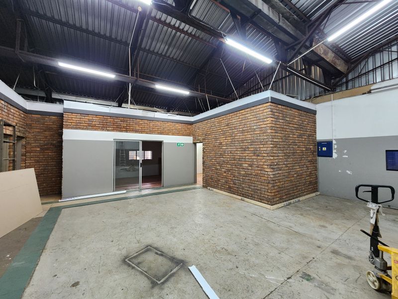 Industrial utility space featuring exposed brick walls, high-clearance corrugated metal roofing, concrete flooring, and fluorescent lighting, suitable for workshop or storage use.