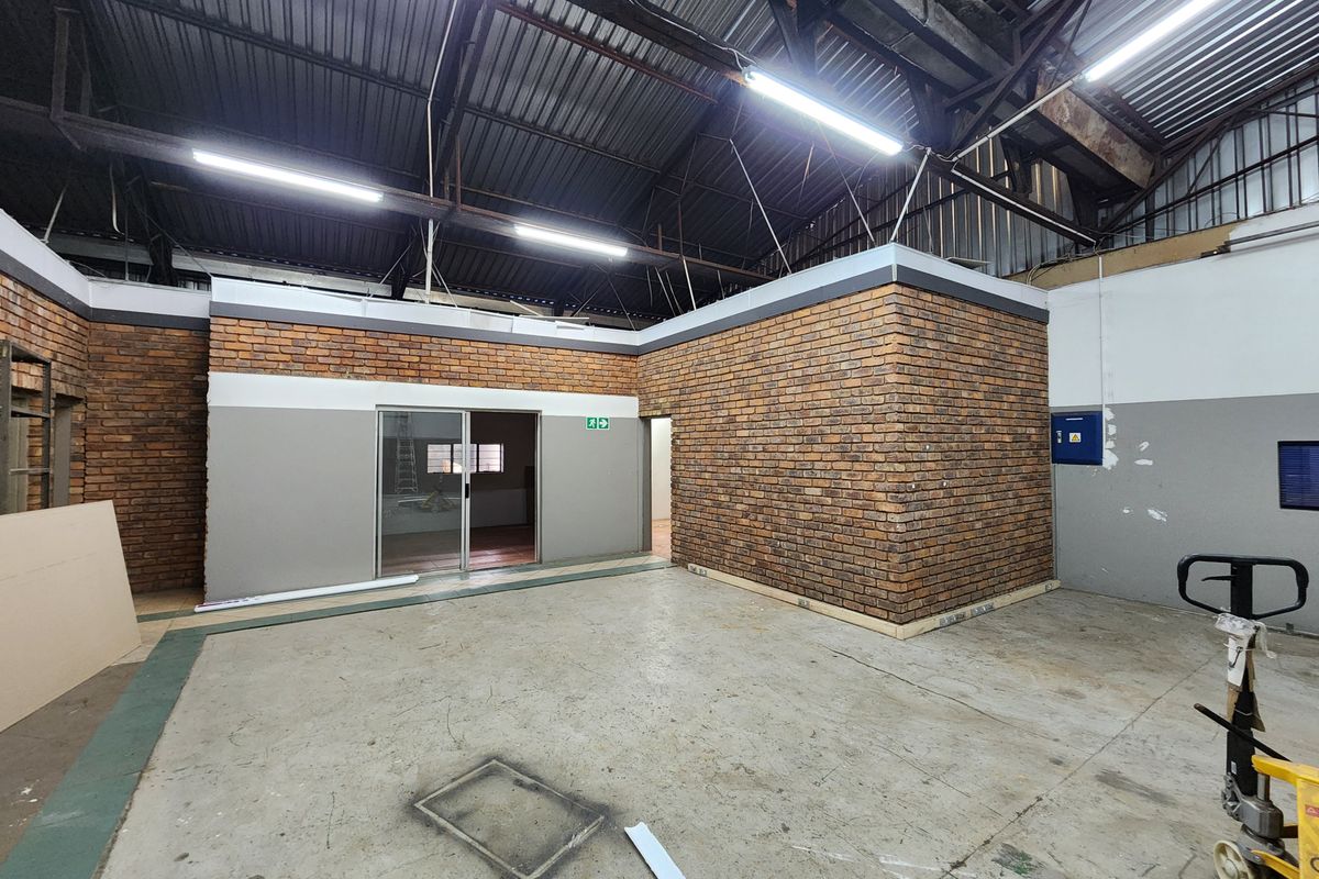 Industrial utility space featuring exposed brick walls, high-clearance corrugated metal roofing, concrete flooring, and fluorescent lighting, suitable for workshop or storage use.