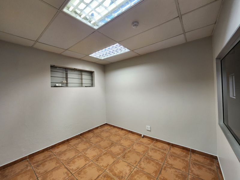 A functional, empty office space featuring ceramic tile flooring, a suspended ceiling with integrated lighting, and a window with security bars.